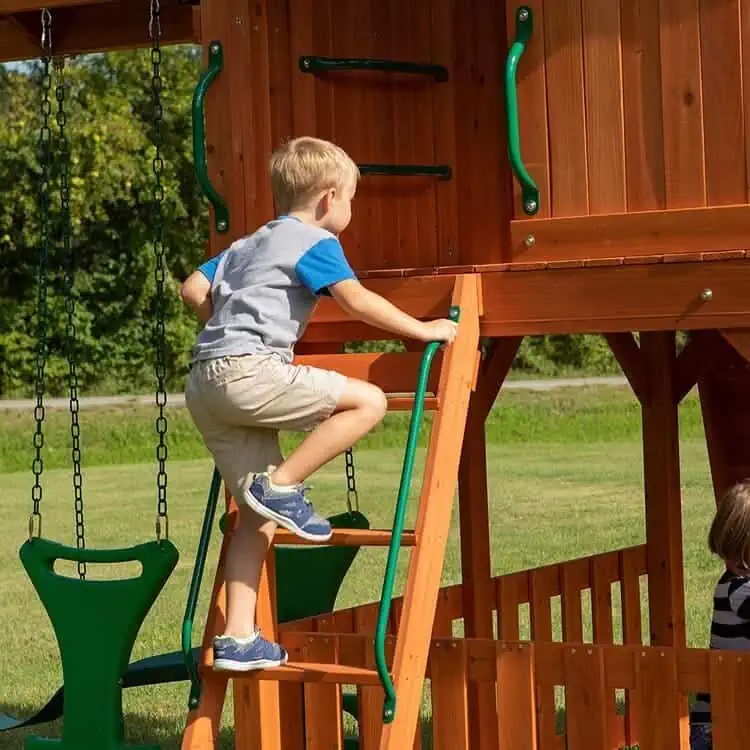 wooden playset with metal safety features, stairs, swings and playhouse, brown and dark green color on green landscape