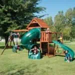 backyard playset with swing set, wooden tower, two slides and a climbing wall, wooden and plastic with green and brown color, on a green landscape