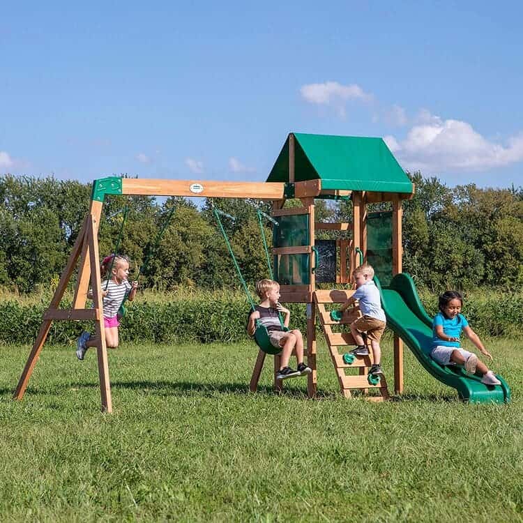 Wooden cedar swing set with covered deck, fort with Mesh panels, rock wall ladder, belt swings with rope chains, slide and chalk board, brown and green color on a green landscape