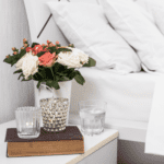 fresh flowers in vase, candle, and glass of water on nightstand next to bed