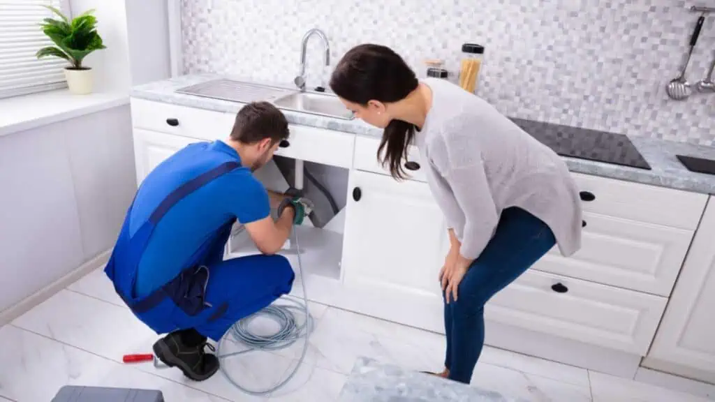 Woman Looking At Male Plumber Cleaning Clogged Sink Pipe With Drained Cable In Kitchen