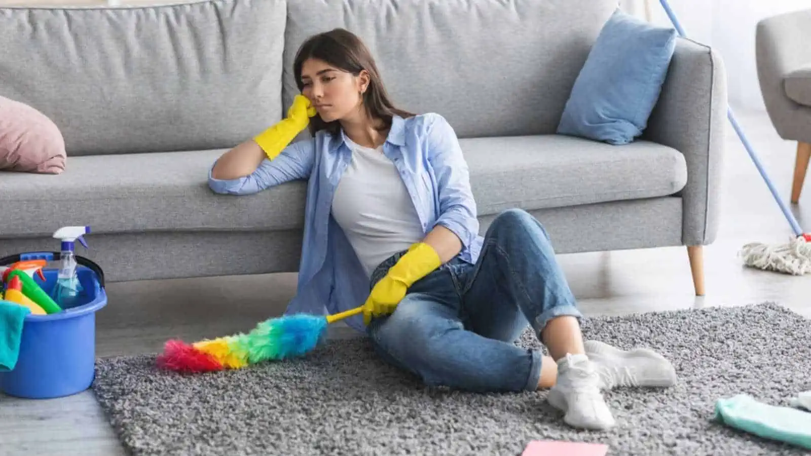 Tired woman leaning on couch, wearing protective gloves, sitting on rug floor carpet surrounded by dust cloth and detergents, taking break while cleaning apartment. Exhausted female looking at mess