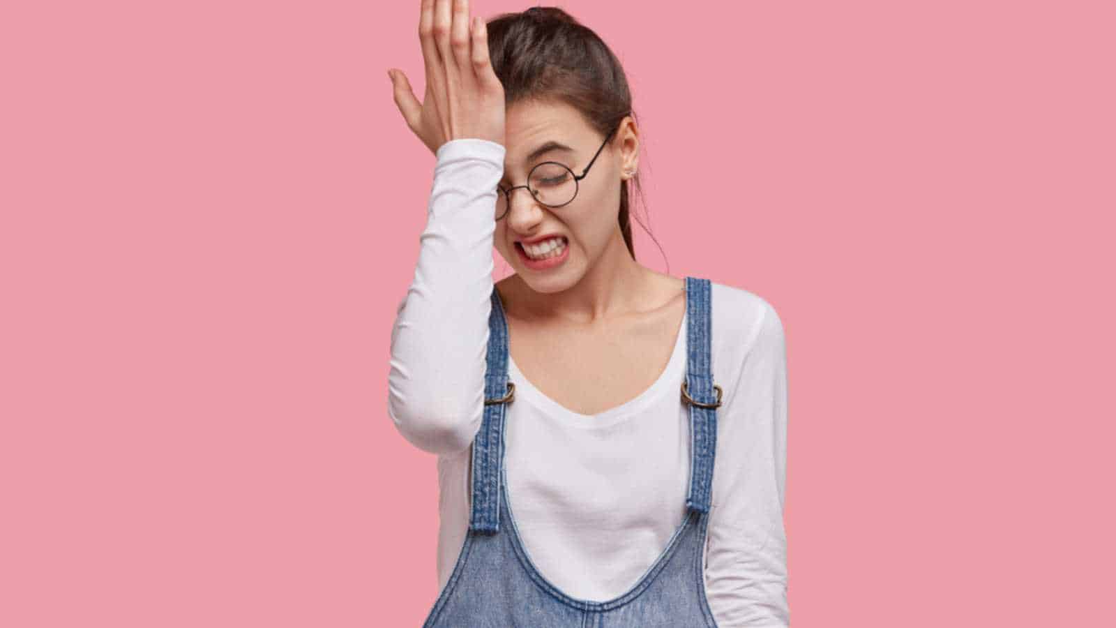 Photo of dissatisfied young woman regrets wrong doing, keeps hand on forehead, clenches teeth, dressed in fashionable outfit, isolated over pink studio wall. Lady forgets something important