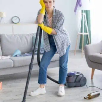 Unhappy young woman standing with vacuum cleaner in messy flat after party, blank space. Upset millennial housewife at dirty room after celebration, garbage scattered everywhere