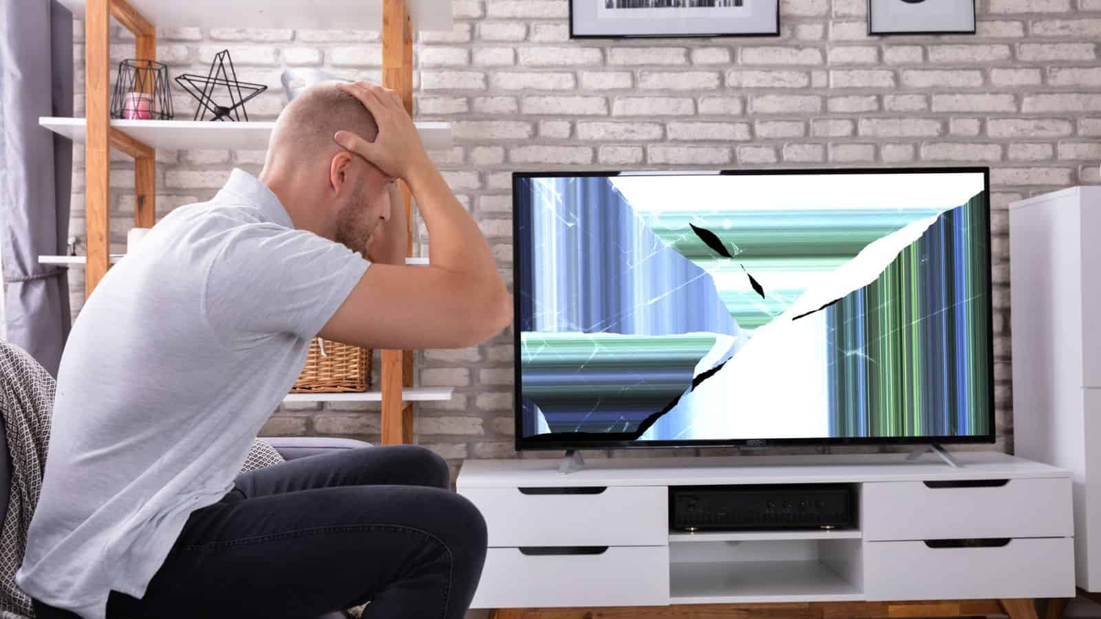 man with hands on head sitting in front of broken tv