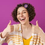 Portrait of an attractive happy young woman with curly brunette hair standing isolated over violet background, thumbs up